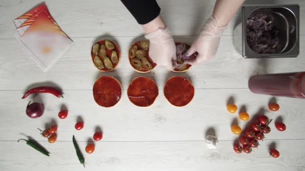 Vista superior de chef profesional cocina una deliciosa hamburguesas en el restaurante de comida rápida. Cocinera en guantes pone cebolla en vinagre en tres panes para hamburguesas — Vídeos de Stock