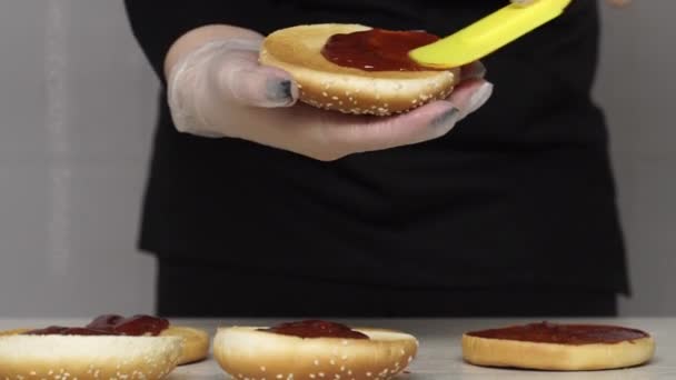 Primer plano de cocineros profesionales una deliciosa hamburguesas en el restaurante de comida rápida. Cocinera en guantes pone ketchup en tres panes asados para hamburguesas — Vídeos de Stock