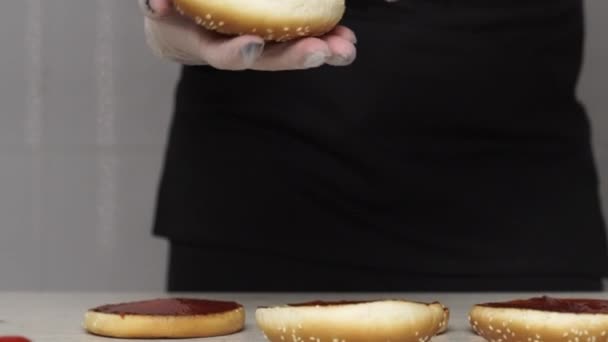 Profesyonel aşçıya yakın çekim, fast food restoranında lezzetli bir burger pişirir. Eldivenli kadın aşçı hamburgerler için üç gül ekmeğine ketçap koyar. — Stok video