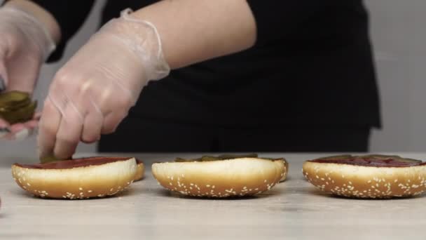 A vista superior do chef profissional cozinha um delicioso hambúrguer no restaurante fast food. Chef feminino em luvas coloca picles em três pães para hambúrgueres — Vídeo de Stock