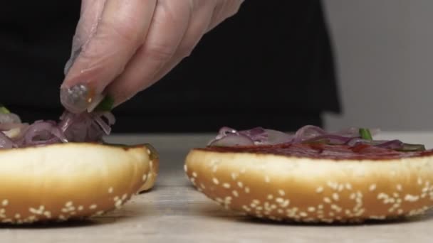 Top view of professional chef cooks a delicious burgers at fast food restaurant. Female chef in gloves puts pickled onion on three breads for burgers — Stock Video