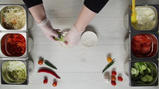 Von oben betrachtet kocht ein professioneller Koch im Fast-Food-Restaurant einen leckeren Burger. Köchin in Handschuhen legt Gurken auf Burger — Stockvideo