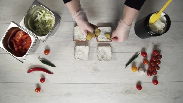 На вид профессиональный повар готовит вкусные бургеры в ресторане быстрого питания. Шеф-повар в перчатках кладет огурцы в бургеры — стоковое видео