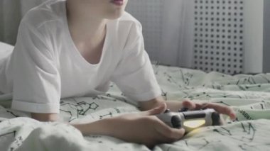 Beyaz tişörtlü genç bir çocuk yatağa uzanıyor ve TV 'de bir oyun platformuyla oyun oynuyor.