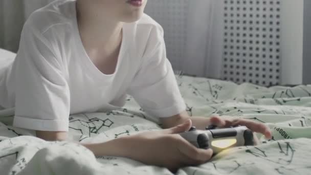 Anak muda berbaju putih berbaring di tempat tidur dan bermain video game di TV dengan gamepad — Stok Video