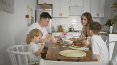 Mutlu bir aile mutfak masasında oturmuş çilek yiyor ve milkshake içiyor.