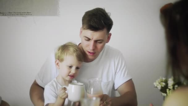 Feliz padre guapo e hijo pequeño se sienta en las rodillas de los padres desayunando por la mañana — Vídeos de Stock