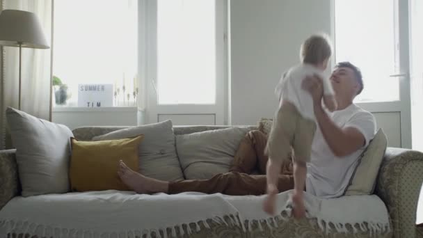 Любящий папа поднимает милый маленький ребенок сын играть самолет весело на диване в гостиной, счастливая семья взрослый отец держит маленький мальчик летит самолет в папочке — стоковое видео