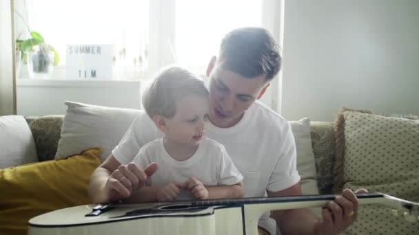 Joven padre enseñando a su pequeño hijo talentoso tocar la guitarra mientras está sentado en la cama en el apartamento de luz . — Vídeos de Stock
