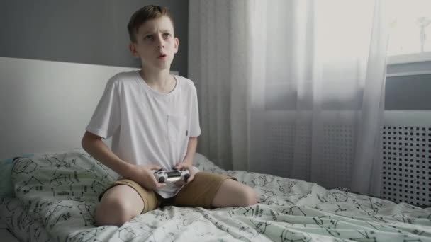 Jovem pré-adolescente concentrado sentado na cama e jogando console de vídeo game — Vídeo de Stock