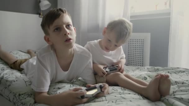Twee kleine broertjes in een wit shirt die in bed zitten en videospelletjes spelen op tv met een gamepad — Stockvideo