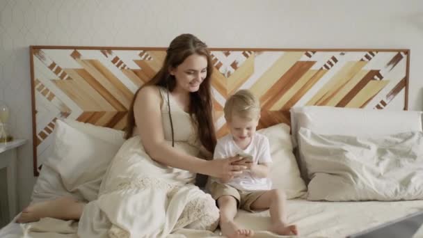Joven madre se sienta en la cama con su hijo y le enseña a usar el teléfono — Vídeos de Stock