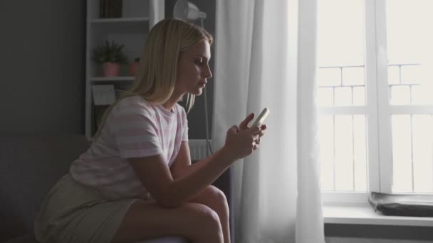 Jovem relaxada feliz segurando telefone inteligente olhando para a tela do celular, aproveitando o uso de aplicativos móveis para fazer compras ou conversar nas mídias sociais sentar no sofá em casa — Vídeo de Stock