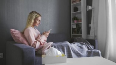 Mutlu ve rahat genç bir kadın cep telefonu ekranına bakıp gülüyor alışveriş için mobil uygulamalar kullanıyor oyun oynuyor sohbet ediyor sosyal medyada sohbet ediyor evde kanepede oturuyor.