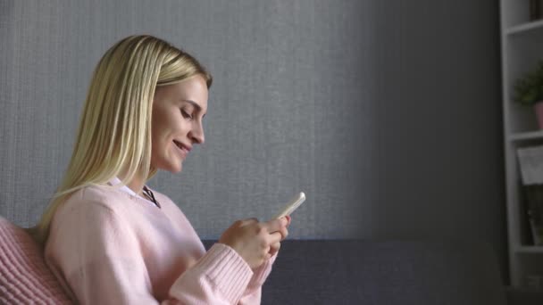 Hermosa joven en el suéter rosa sosteniendo un teléfono inteligente en las manos y escribiendo en su pantalla mientras charla en la sala de estar en casa — Vídeo de stock