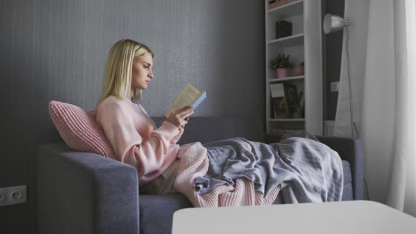 Blanke aantrekkelijke vrouw die een boek leest op de grijze bank in de gezellige woonkamer — Stockvideo