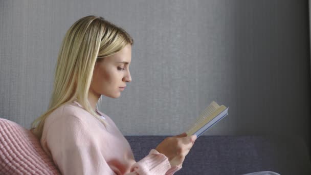 Primer plano de mujer atractiva caucásica leyendo un libro en el sofá gris en la acogedora sala de estar — Vídeo de stock