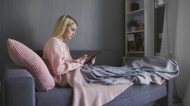Çekici, gülümseyen beyaz kız kanepede oturuyor ve tablet kullanıyor. Sabahları sosyal medyayı kullanan neşeli genç bir kadının portresi