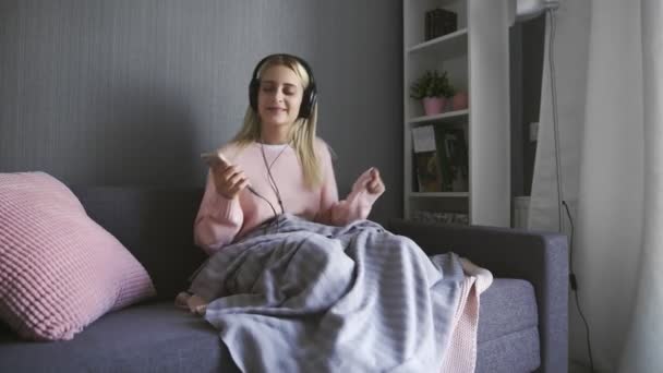 Junge Frau mit Kopfhörern hört fröhliche Musik und tanzt auf Couch — Stockvideo