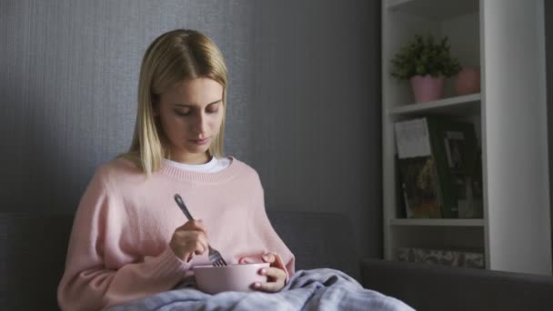 Молодая женщина сидит на диване и держит миску с салатом — стоковое видео