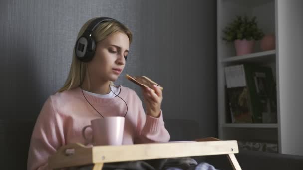 Jovem mulher senta-se no sofá come torrada com pasta de chocolate e ouvir a música no smartphone — Vídeo de Stock