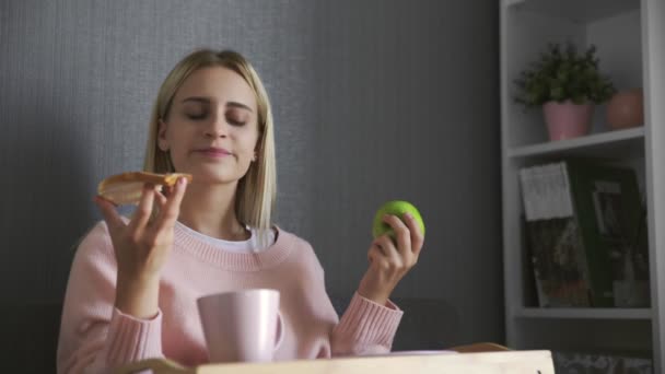 Mujer joven se sienta en el sofá y hace una elección qué comer tostadas con chocolate o manzana — Vídeos de Stock