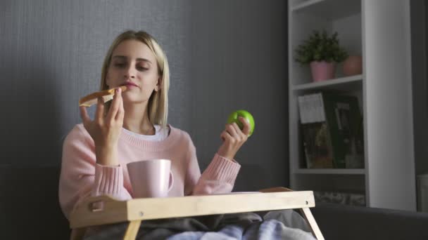 Mladá žena sedí na gauči a dělá výběr, co jíst toast s čokoládou nebo jablkem — Stock video