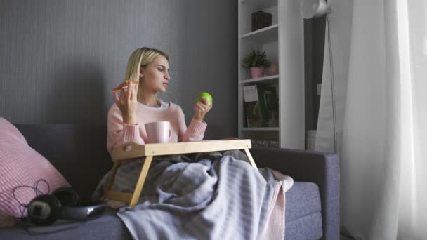 Mladá žena sedí na gauči a dělá výběr, co jíst toast s čokoládou nebo jablkem — Stock video