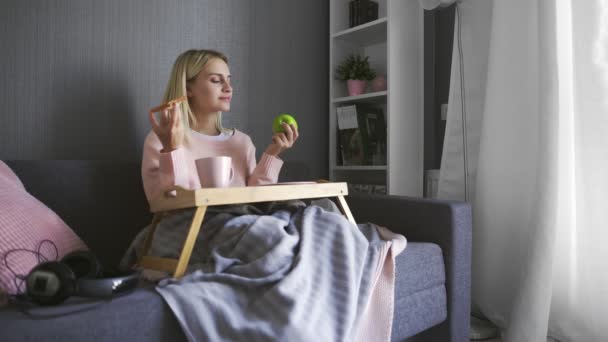 Young woman sits on couch and makes a choice what to eat toast with chocolate or apple — Stock Video