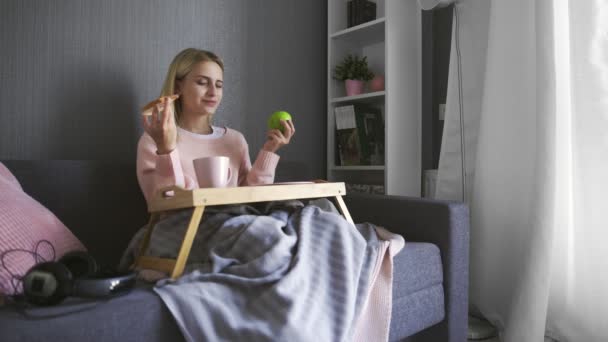 Mujer joven se sienta en el sofá y hace una elección qué comer tostadas con chocolate o manzana — Vídeos de Stock