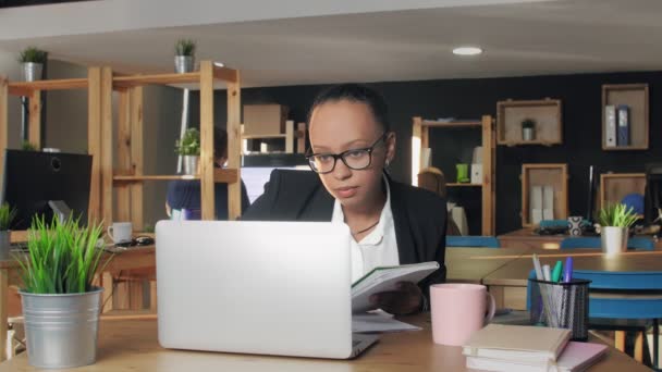 Fiatal afro-amerikai nő gépel laptopon az irodában, aztán jegyzetfüzetbe ír. — Stock videók