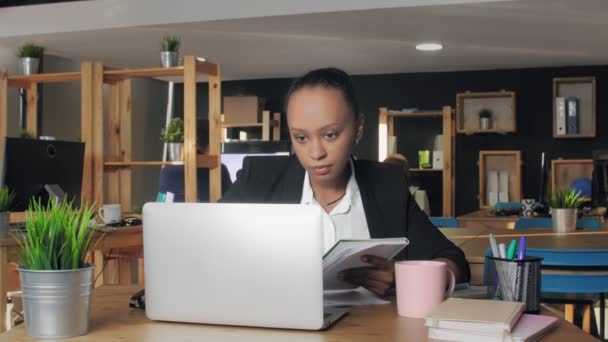 Junge Afroamerikanerin tippt im Büro auf Laptop und schreibt dann in Notizbuch — Stockvideo