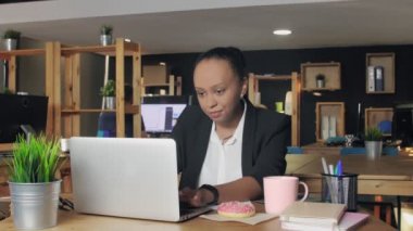 Genç Afrikalı Amerikalı kadın defter üzerinde çalışıyor ve bazen donutuna bakıyor.