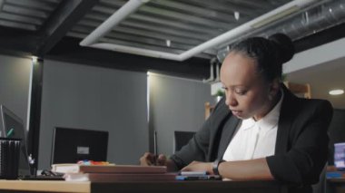 Genç Afro-Amerikan kadının ofiste dizüstü bilgisayarda yazı yazıp sonra da deftere yazışının yan görüntüsü