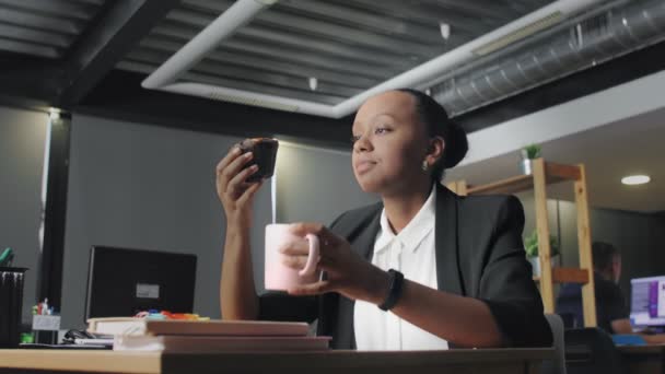 Vista de la triste mujer afroamericana almorzando en la oficina. Mujer come panecillo y bebe un café — Vídeos de Stock