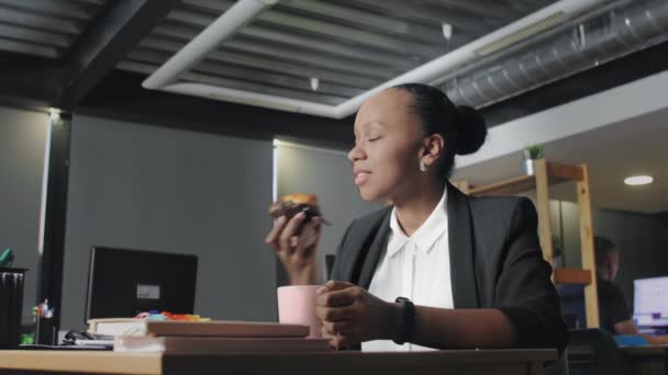 Widok afrykańskiej amerykanki jedzącej lunch w biurze. Kobieta je muffinki i pije kawę. — Wideo stockowe