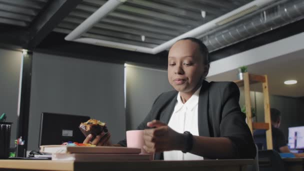 Widok afrykańskiej amerykanki jedzącej lunch w biurze. Kobieta je muffinki i pije kawę. — Wideo stockowe