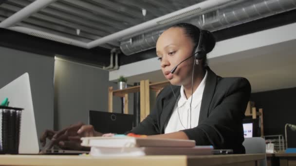 Eine junge Afroamerikanerin mit Headset spricht mit einer Kundin, während sie auf ihren Computermonitor blickt. — Stockvideo