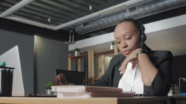 Vue vers le bas de femme afro-américaine ennuyeuse avec casque parle à un client tout en regardant son écran d'ordinateur . — Video