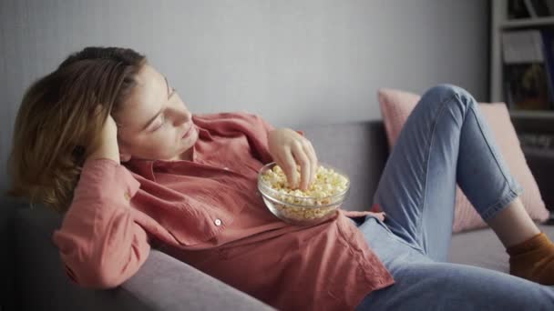 Vista lateral da jovem mulher sentada no sofá, comendo pipocas e assistindo TV — Vídeo de Stock