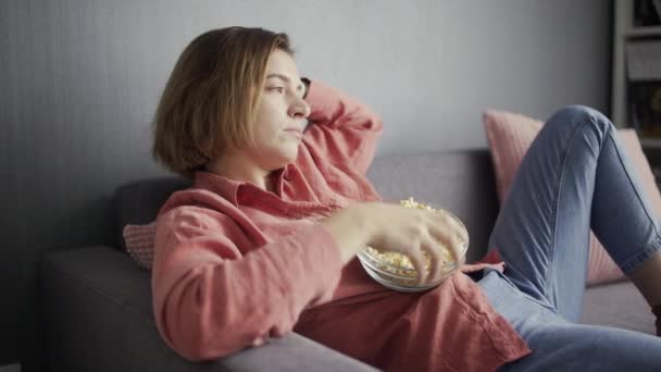Ленивая женщина сидит на диване, ест попкорн и смотрит телевизор — стоковое видео
