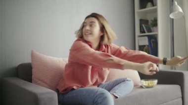 Genç, heyecanlı bir kadın kanepede oturuyor, arkadaşıyla konuşuyor ve TV seyrediyor.