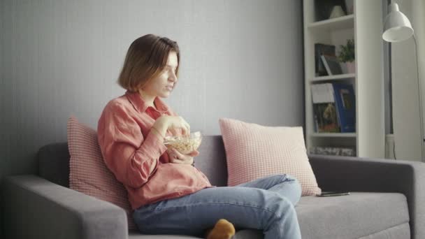 Молодая сконцентрированная женщина сидит на диване, ест попкорн и смотрит телевизор. — стоковое видео