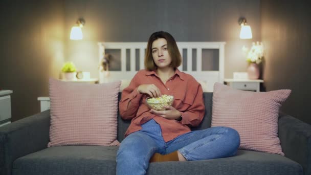 Mujer joven sentada en el sofá, comiendo palomitas de maíz y viendo la televisión — Vídeo de stock
