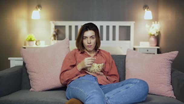 Молодая женщина сидит на диване, ест попкорн, смотрит телевизор и падает снова — стоковое видео