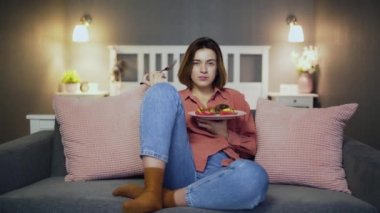 Genç bir kadın TV 'nin önündeki kanepede yemek yiyor..