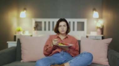 Genç bir kadın TV 'nin önündeki kanepede yemek yiyor..