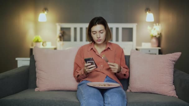 Mujer joven cenando en el sofá y usando un smartphone — Vídeos de Stock