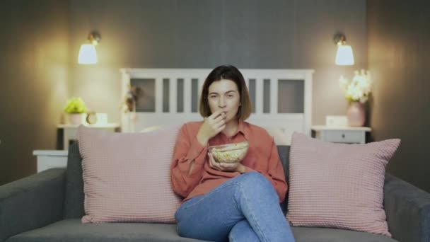 Glückliche junge Frau sitzt auf dem Sofa, isst Popcorn, sieht fern und lacht — Stockvideo