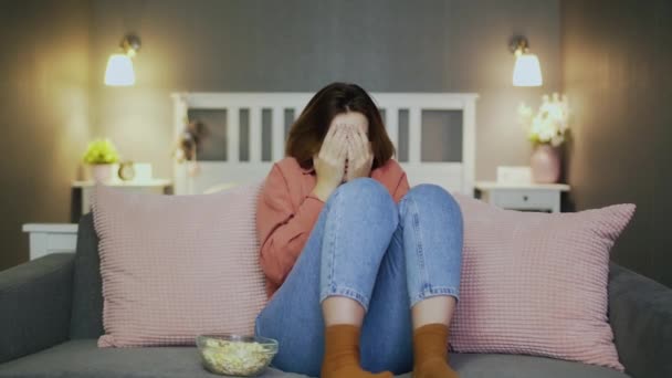 Испуганная молодая женщина сидит на диване, ест попкорн, смотрит телевизор и закрывает лицо руками — стоковое видео